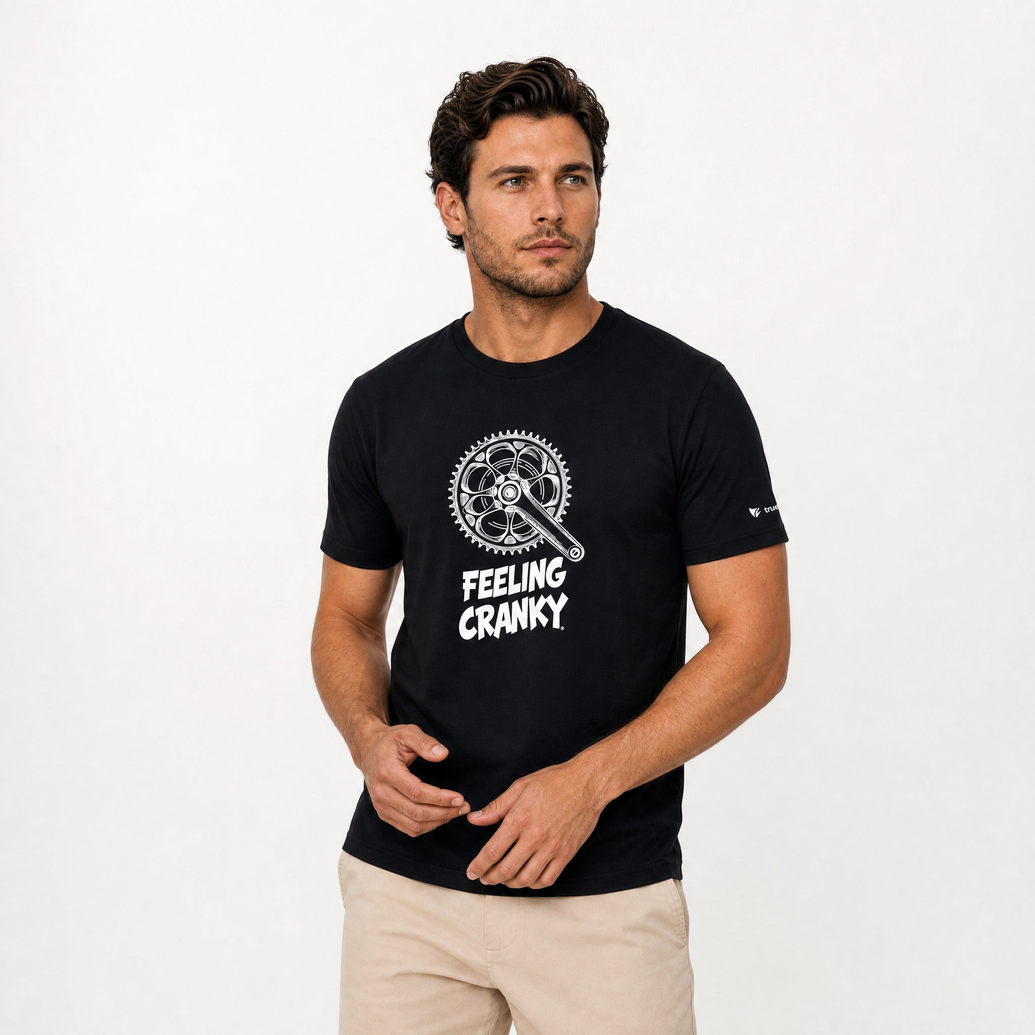 A man wearing a black short-sleeve t-shirt with a white graphic of a bicycle chainring and a wrench, with the text 'FEELING CRANKY' printed below it. He is also wearing beige pants, set against a plain white studio background.