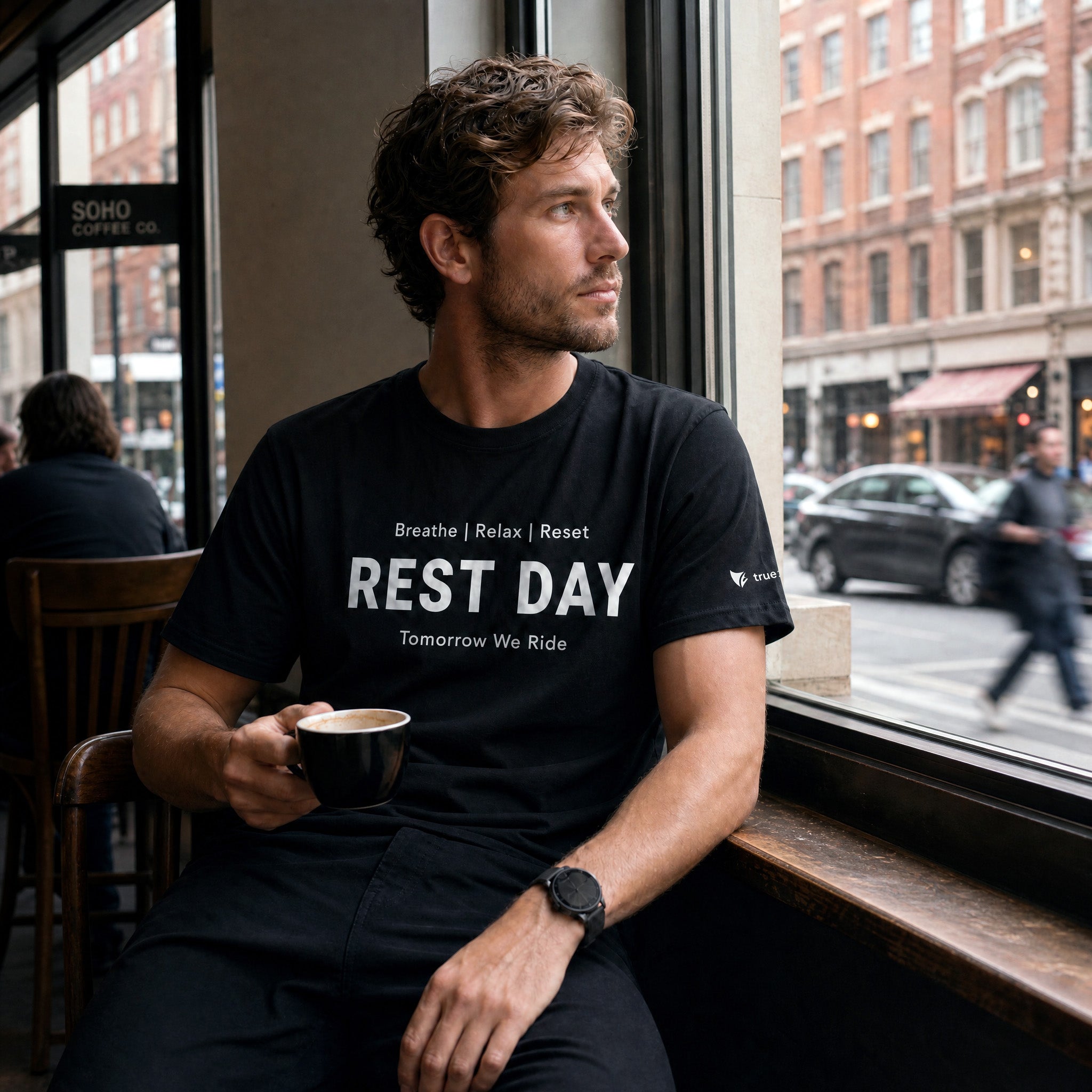 Man sitting in a cafe wearing a black t-shirt with 'REST DAY' text, holding a coffee cup.