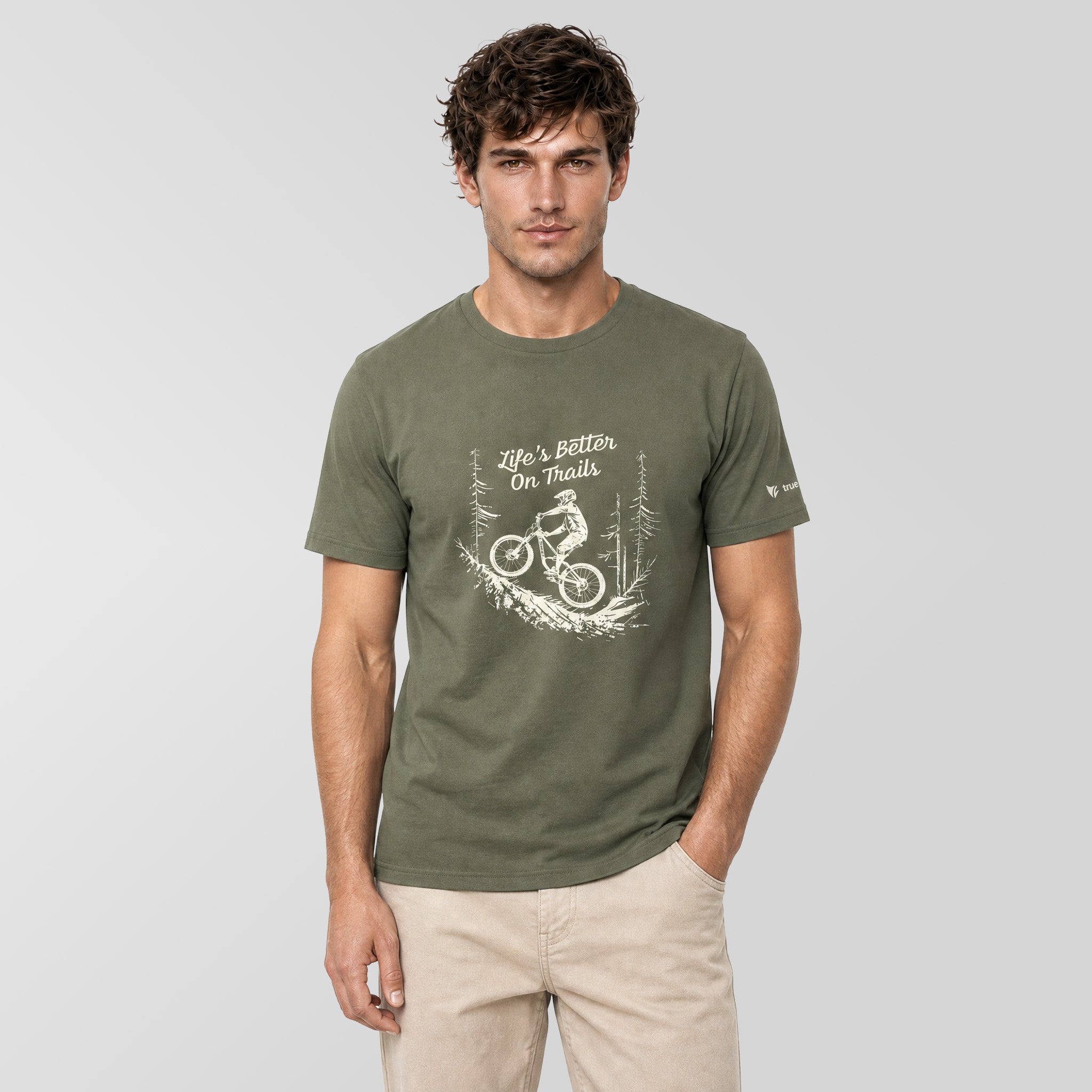 A man wearing an olive green t-shirt with a white graphic of a mountain biker and the text ‘life’s Better On Trails' on the front, paired with beige pants, against a solid light gray background.