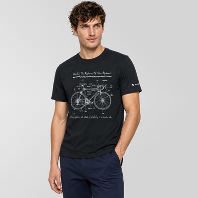 A man wearing a black short-sleeve t-shirt with a white bicycle patent drawing and text on the front, paired with navy pants, against a solid gray background.