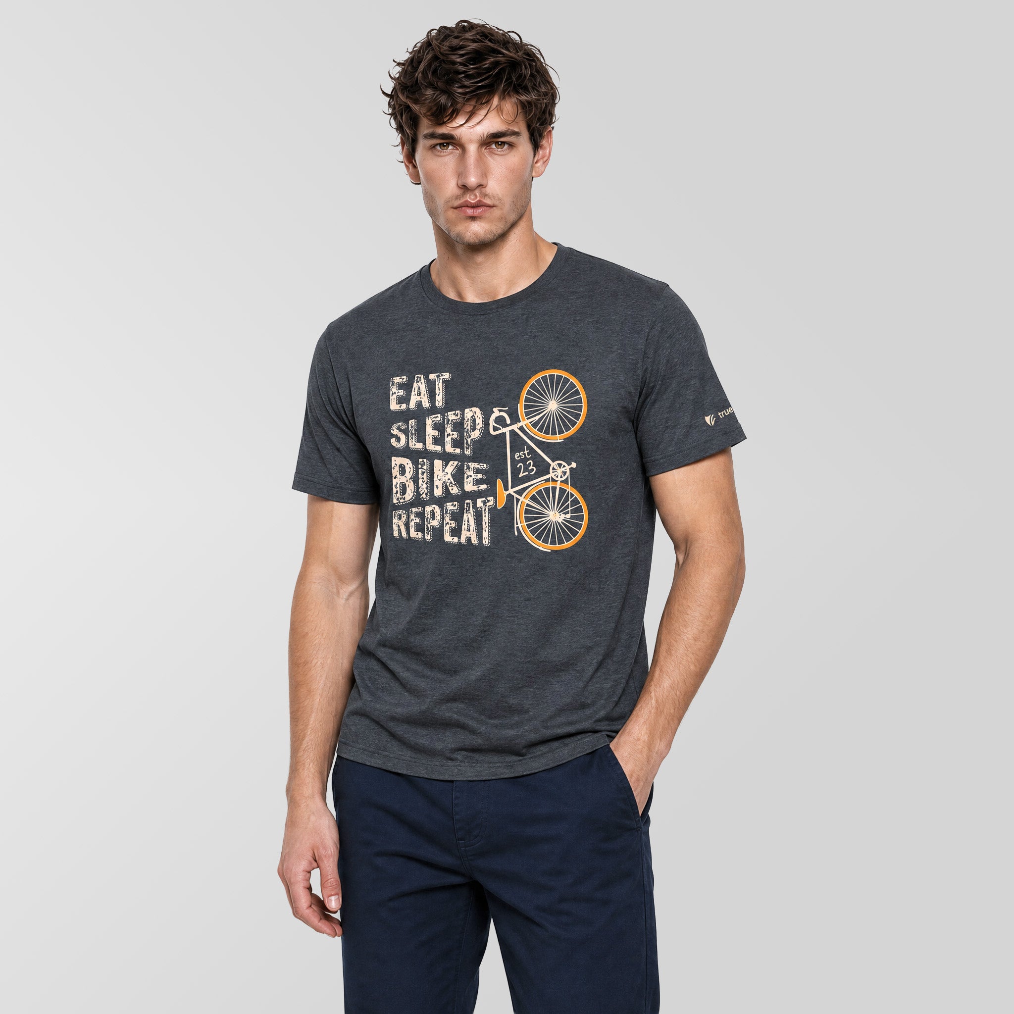 A man wearing a dark gray heather graphic t-shirt with a bicycle and 'EAT SLEEP & BIKE, REPEAT!' print, paired with navy blue pants, against a light gray studio background.