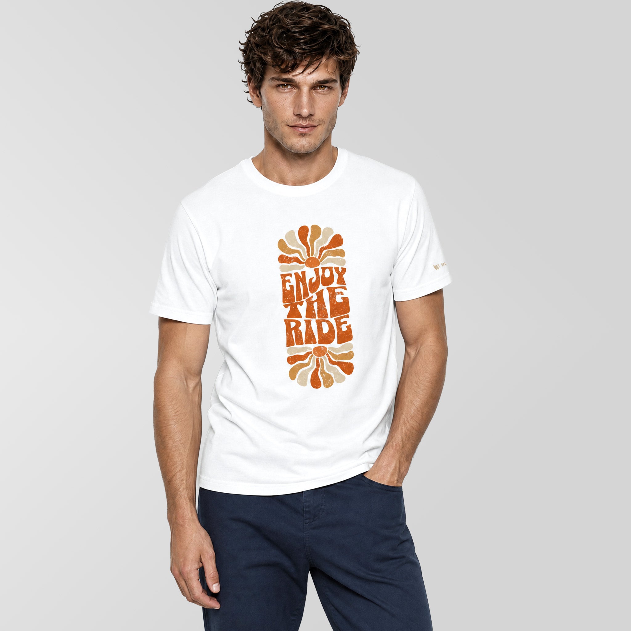 A man wearing a white crewneck t-shirt with a retro orange and yellow 'ENJOY THE RIDE' graphic on the front, paired with navy blue pants, against a solid gray background.