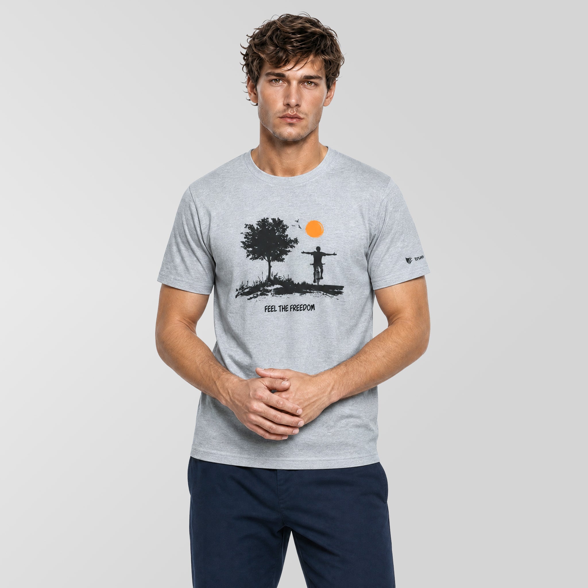A man wearing a light gray crewneck t-shirt with a black graphic of a tree, a figure on a cross, and an orange sun, paired with navy blue pants, against a light gray studio background.