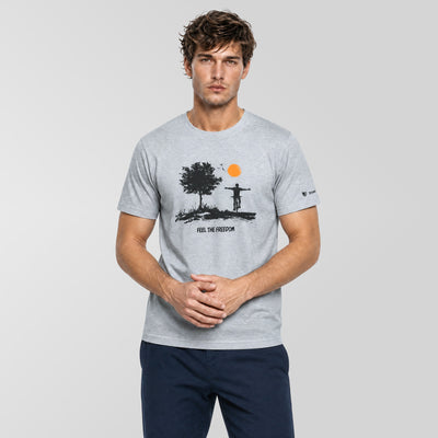 A man wearing a light gray crewneck t-shirt with a black graphic of a tree, a figure on a cross, and an orange sun, paired with navy blue pants, against a light gray studio background.