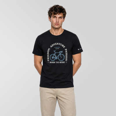 A man modeling a black short-sleeve t-shirt with a blue bicycle graphic and the text 'FREEDOM ADVENTURE PASSION BORN TO RIDE' on the front, paired with beige pants, against a light gray studio background.