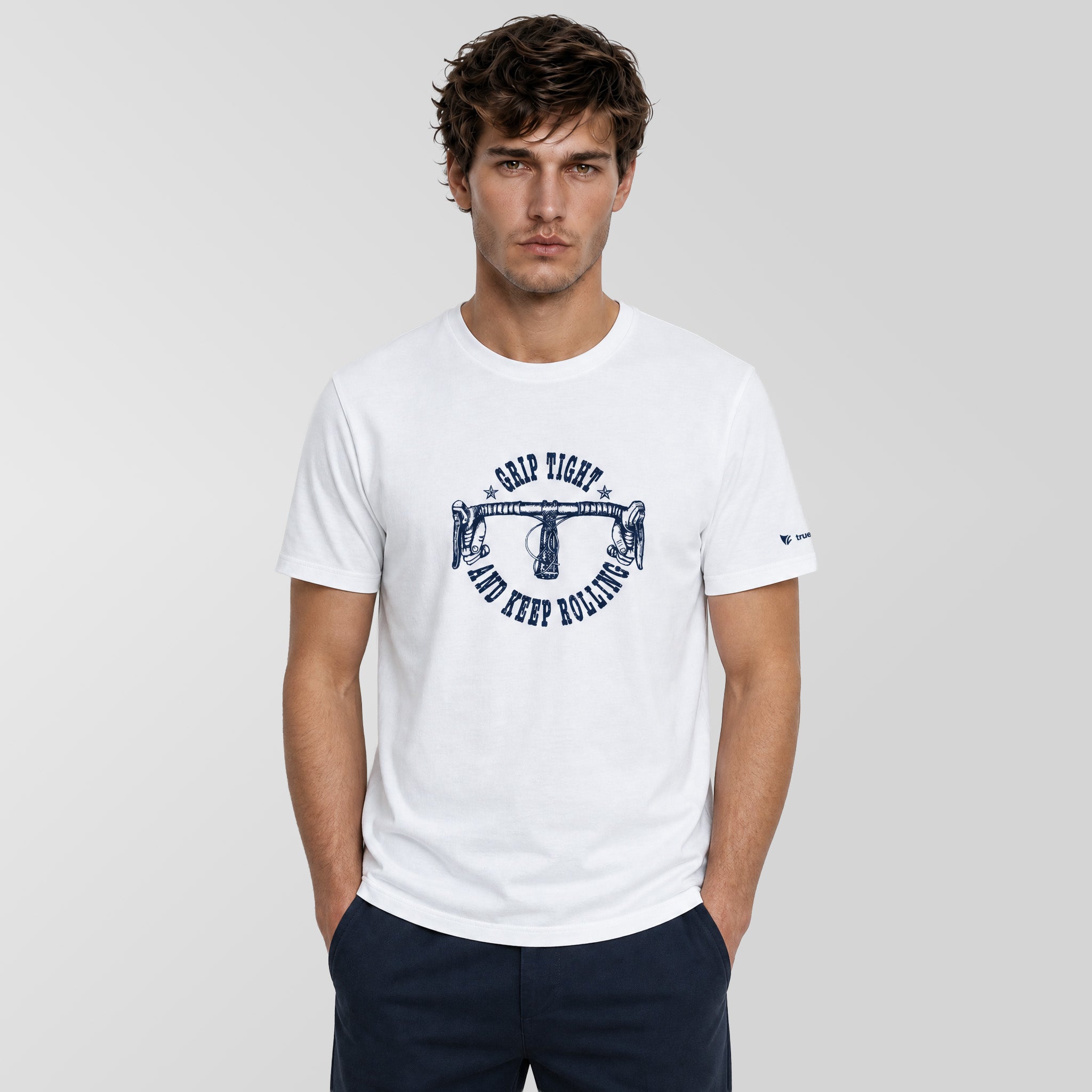 A man wearing a white crew-neck t-shirt with a navy blue graphic print on the front that reads ‘GRIP RIGHT AND KEEP BALANCE' around a balance scale illustration. The background is a clean, light gray studio wall.