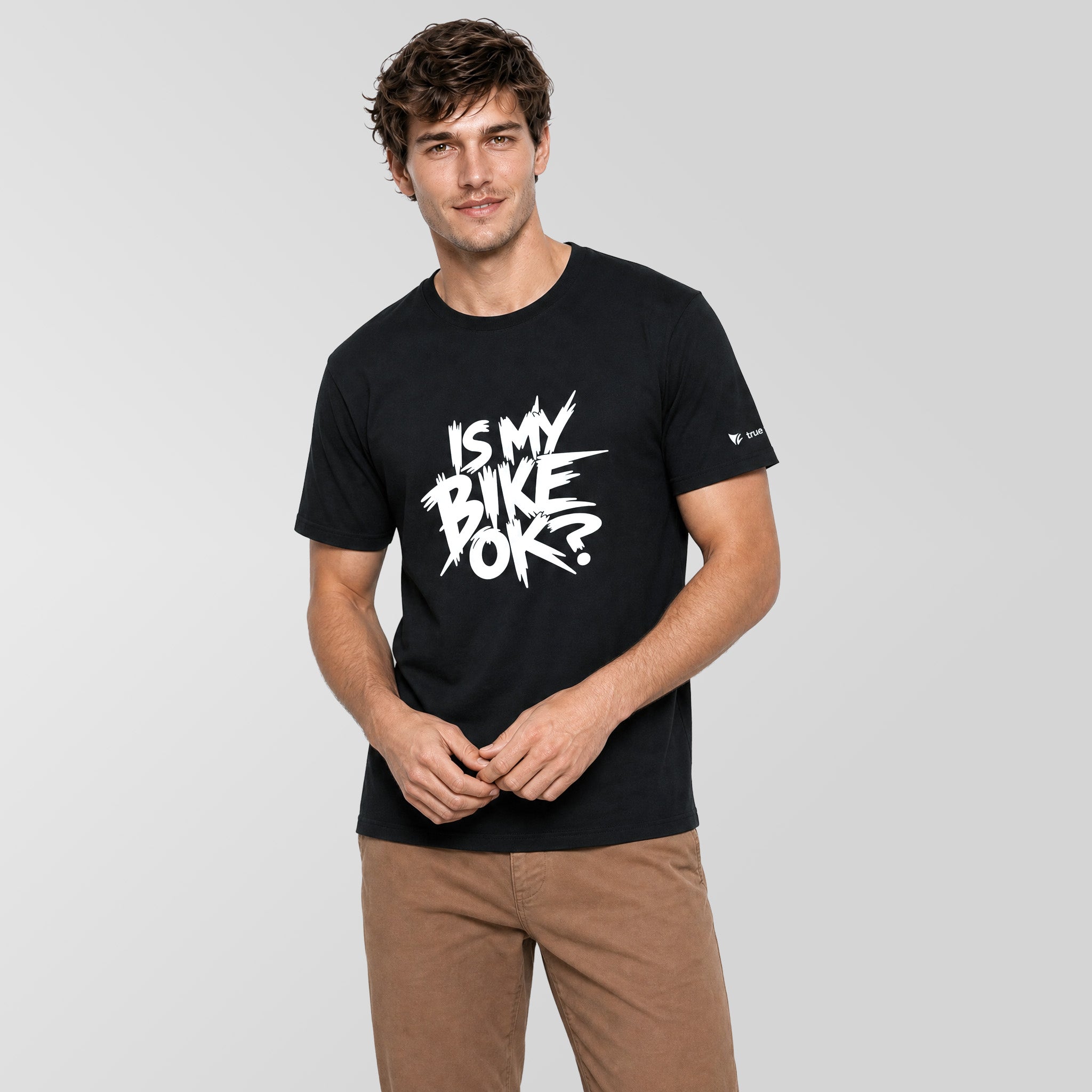A man wearing a black short-sleeve t-shirt with a white graphic text print that reads 'IS MY BIKE OK?' on the chest, paired with brown pants, against a solid light gray background.