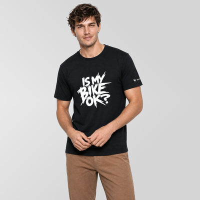 A man wearing a black short-sleeve t-shirt with a white graphic text print that reads 'IS MY BIKE OK?' on the chest, paired with brown pants, against a solid light gray background.