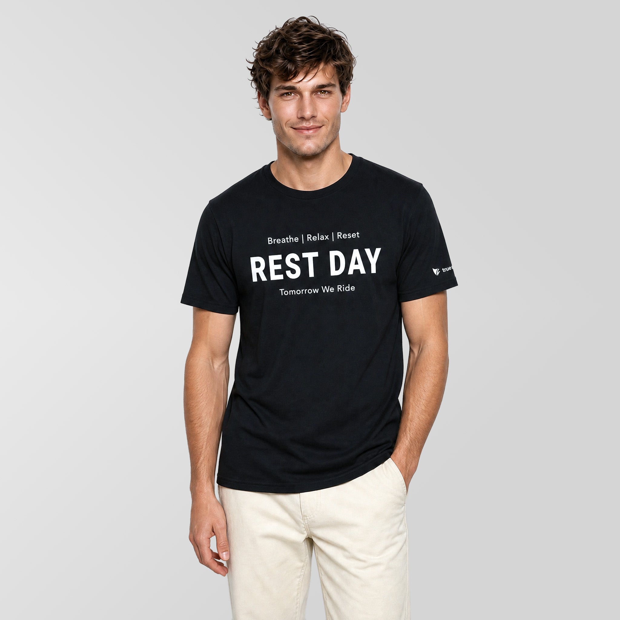 A man wearing a black crewneck t-shirt with white text that reads 'REST DAY' and 'Tomorrow We Rise' on the front, paired with off-white pants, against a solid light gray background.