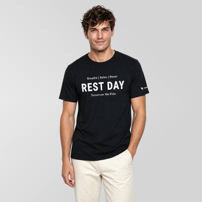 A man wearing a black crewneck t-shirt with white text that reads 'REST DAY' and 'Tomorrow We Rise' on the front, paired with off-white pants, against a solid light gray background.
