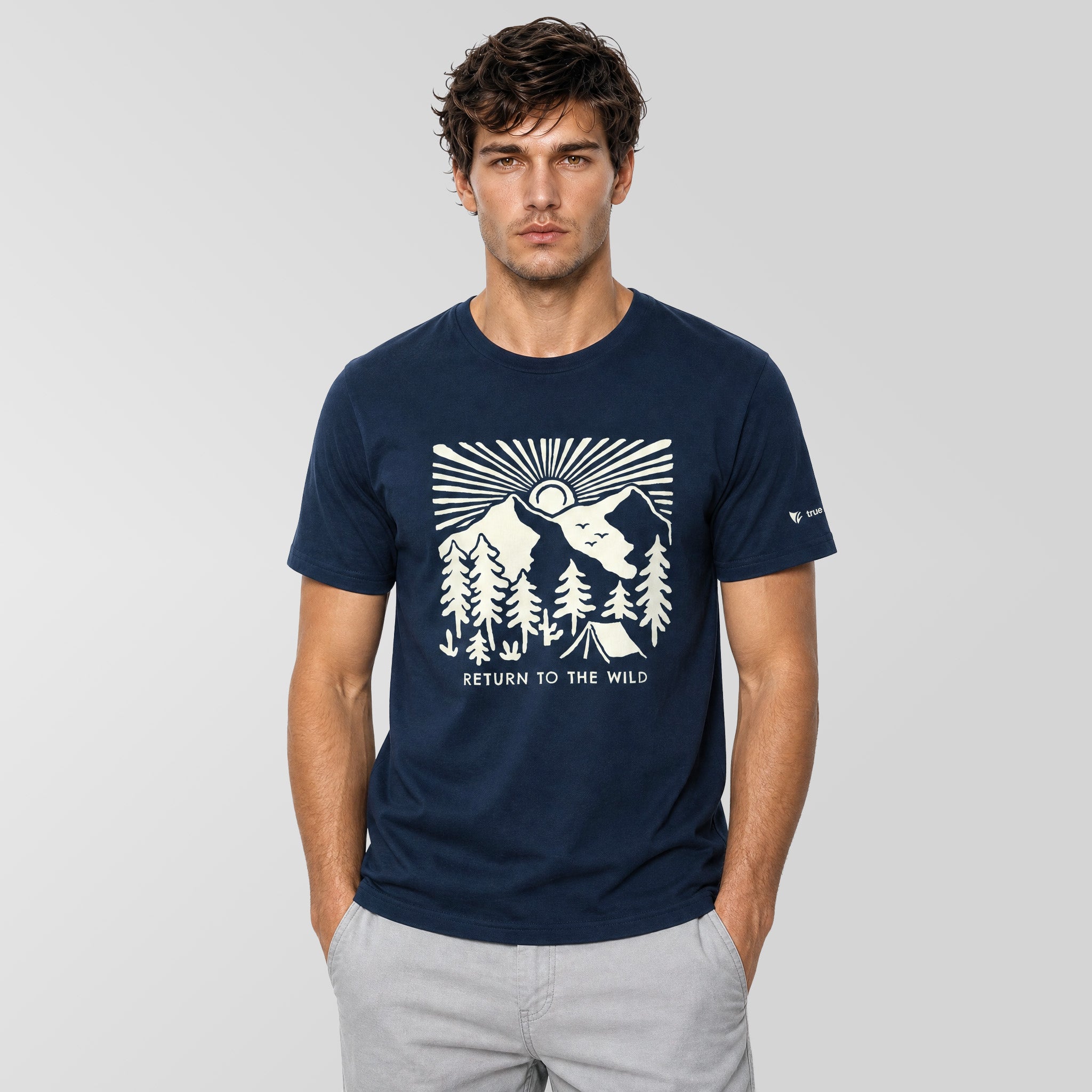 A man wearing a navy blue crewneck t-shirt with a white graphic print of a mountain, trees, and a wolf, with the text 'RETURN TO THE WILD' on the front. He is also wearing light gray pants, standing against a light gray studio background.