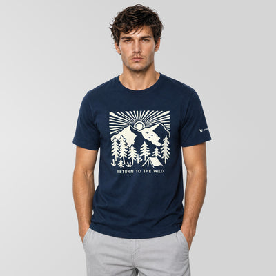 A man wearing a navy blue crewneck t-shirt with a white graphic print of a mountain, trees, and a wolf, with the text 'RETURN TO THE WILD' on the front. He is also wearing light gray pants, standing against a light gray studio background.