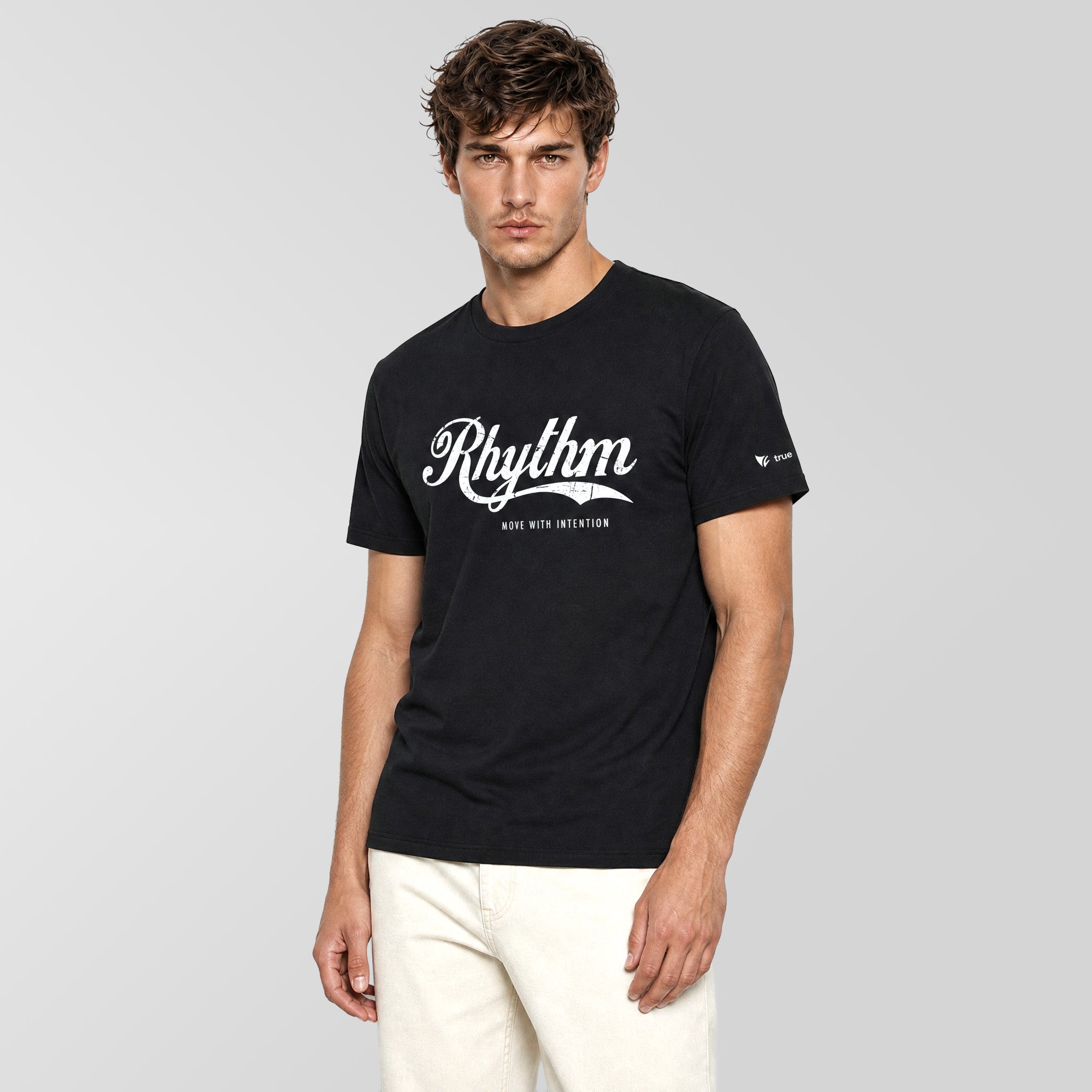 A man wearing a black short-sleeve t-shirt with a white 'Rhythm' graphic on the chest, paired with off-white trousers, standing against a light gray studio background.