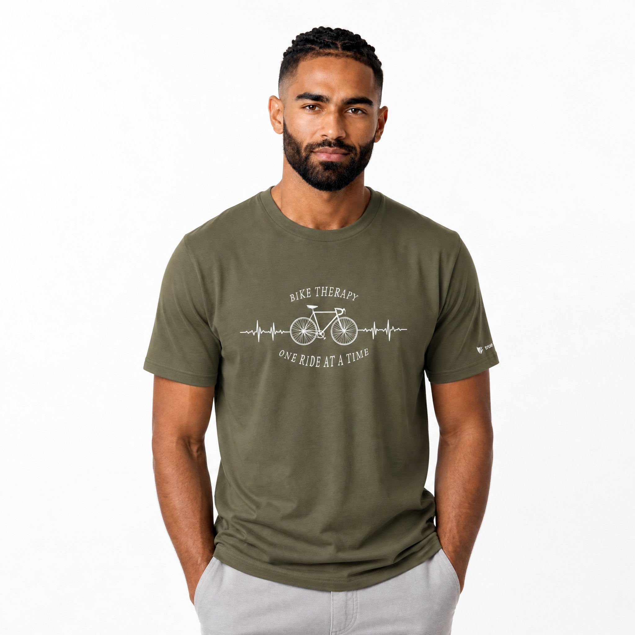 A man wearing an olive green crewneck t-shirt with a white graphic print that reads ‘BIKE THERAPY' and 'ONE RIDE AT A TIME' around a bicycle and EKG line, paired with light gray pants, against a plain white background.