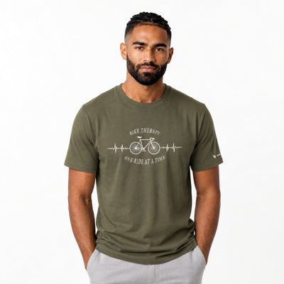 A man wearing an olive green crewneck t-shirt with a white graphic print that reads ‘BIKE THERAPY' and 'ONE RIDE AT A TIME' around a bicycle and EKG line, paired with light gray pants, against a plain white background.