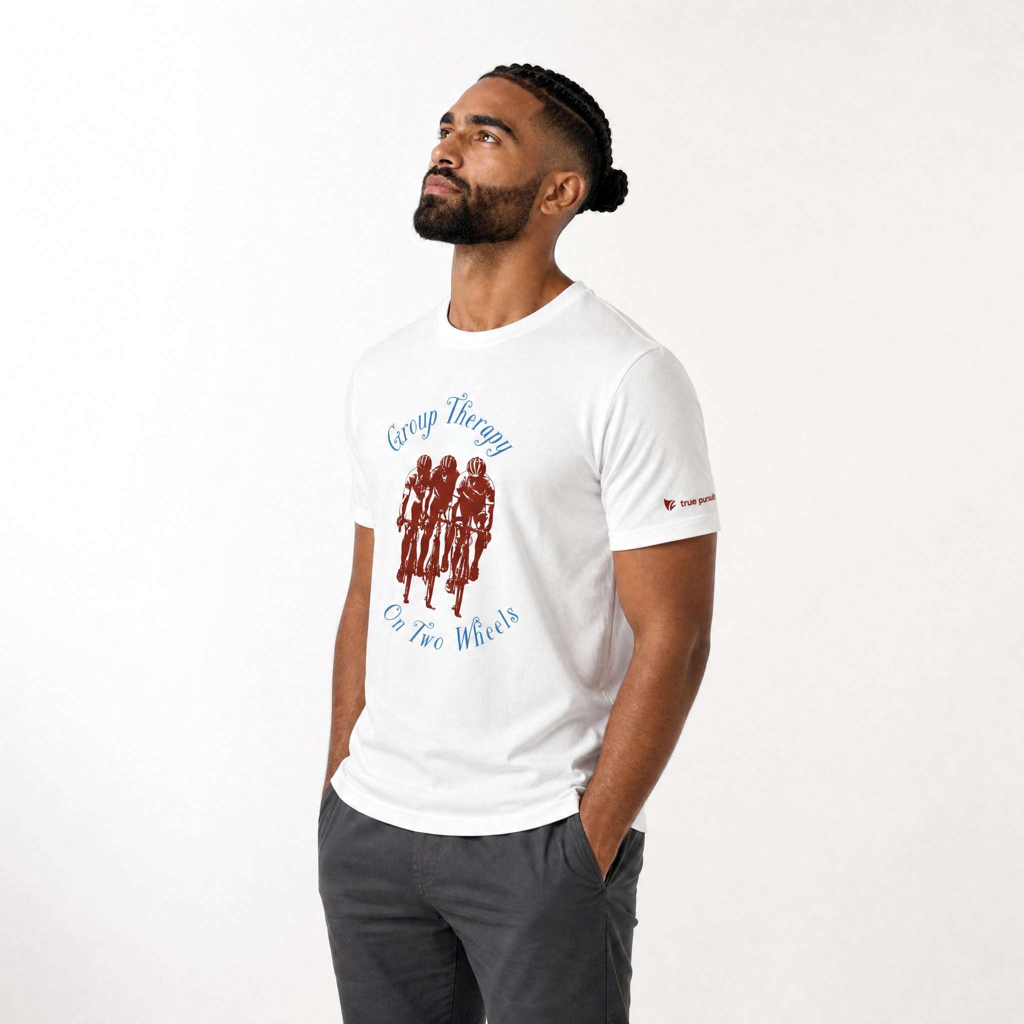 A man wearing a white graphic t-shirt with a red and blue print that reads ‘Group Therapy On Two Wheels' and a small red logo on the sleeve, paired with dark gray pants, against a plain white background.