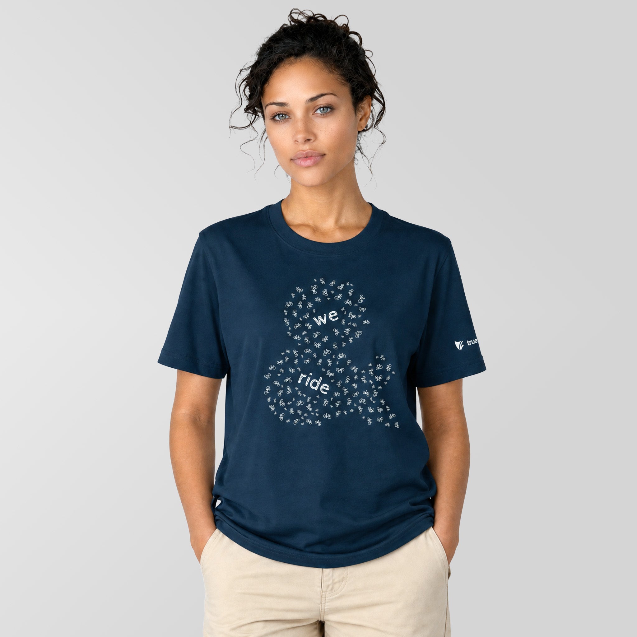 A woman modeling a navy blue crewneck t-shirt with a white graphic of a bicycle made of small hearts and the words 'we ride' on the front, paired with beige pants, against a solid light gray background.