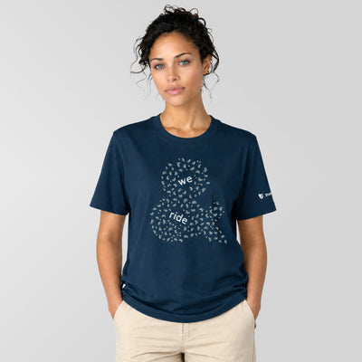 A woman modeling a navy blue crewneck t-shirt with a white graphic of a bicycle made of small hearts and the words 'we ride' on the front, paired with beige pants, against a solid light gray background.