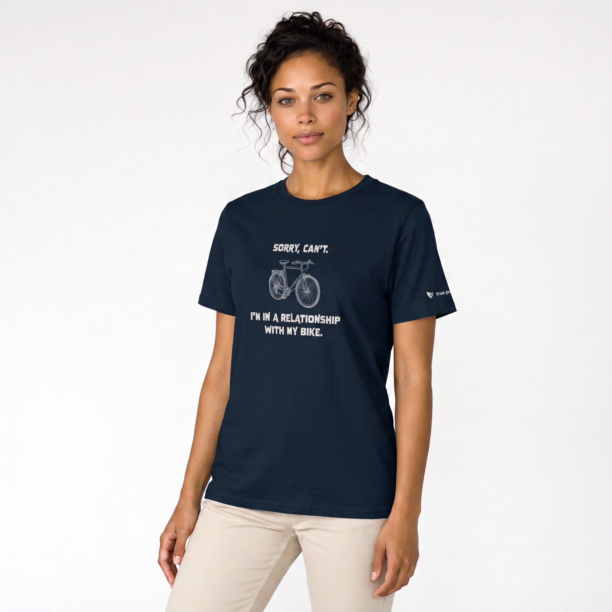 A woman modeling a navy blue crewneck t-shirt with a white graphic of a bicycle and the text 'SORRY CAN'T I' I'M IN A RELATIONSHIP WITH MY BIKE.' on the front, paired with light beige pants against a plain white studio background.