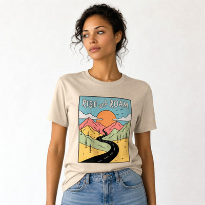 Woman wearing a beige t-shirt with a graphic design and text 'Rise and Roam' on a white background