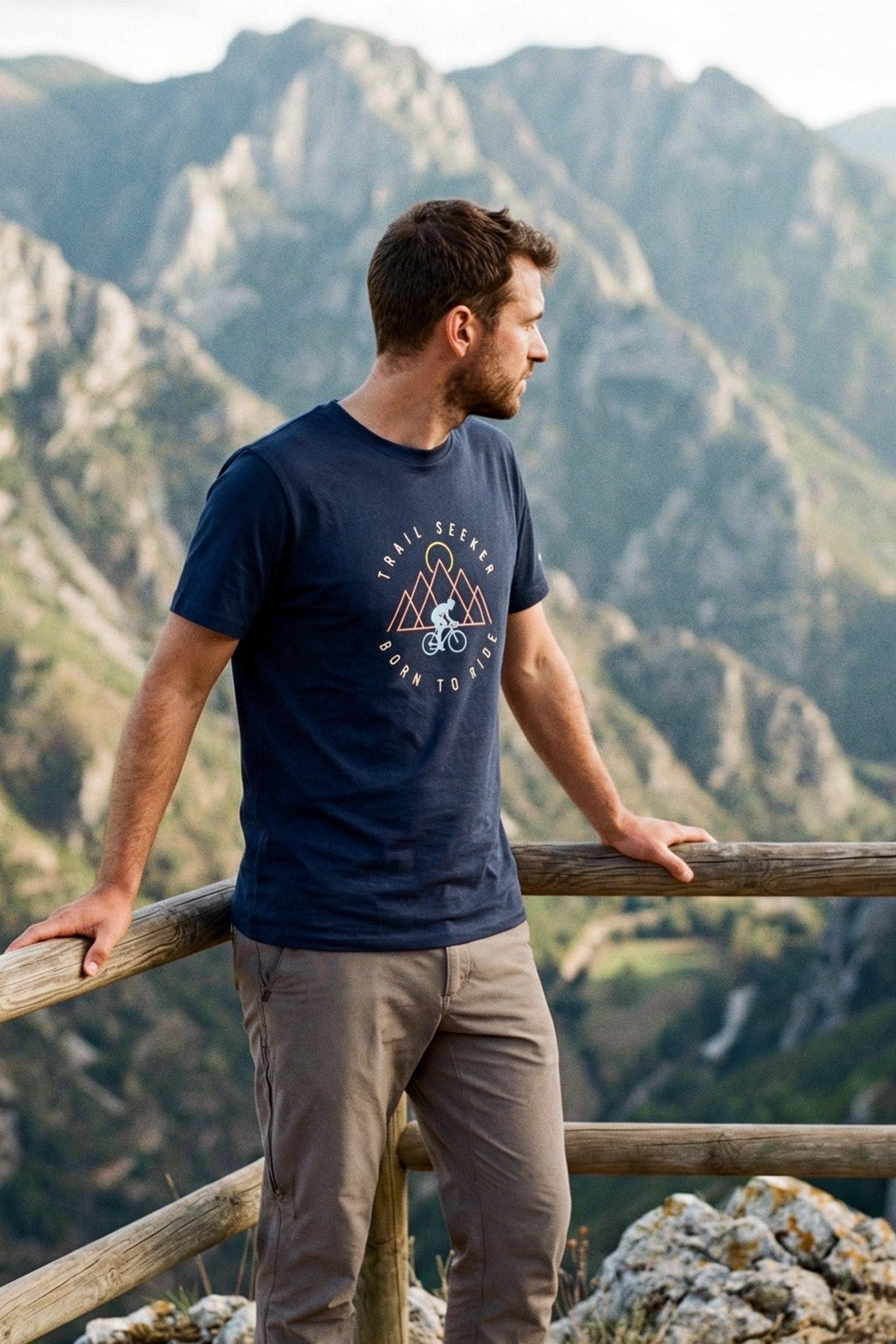 Man wearing a navy blue t-shirt with a mountain graphic, standing on a wooden railing with a mountainous landscape in the background.