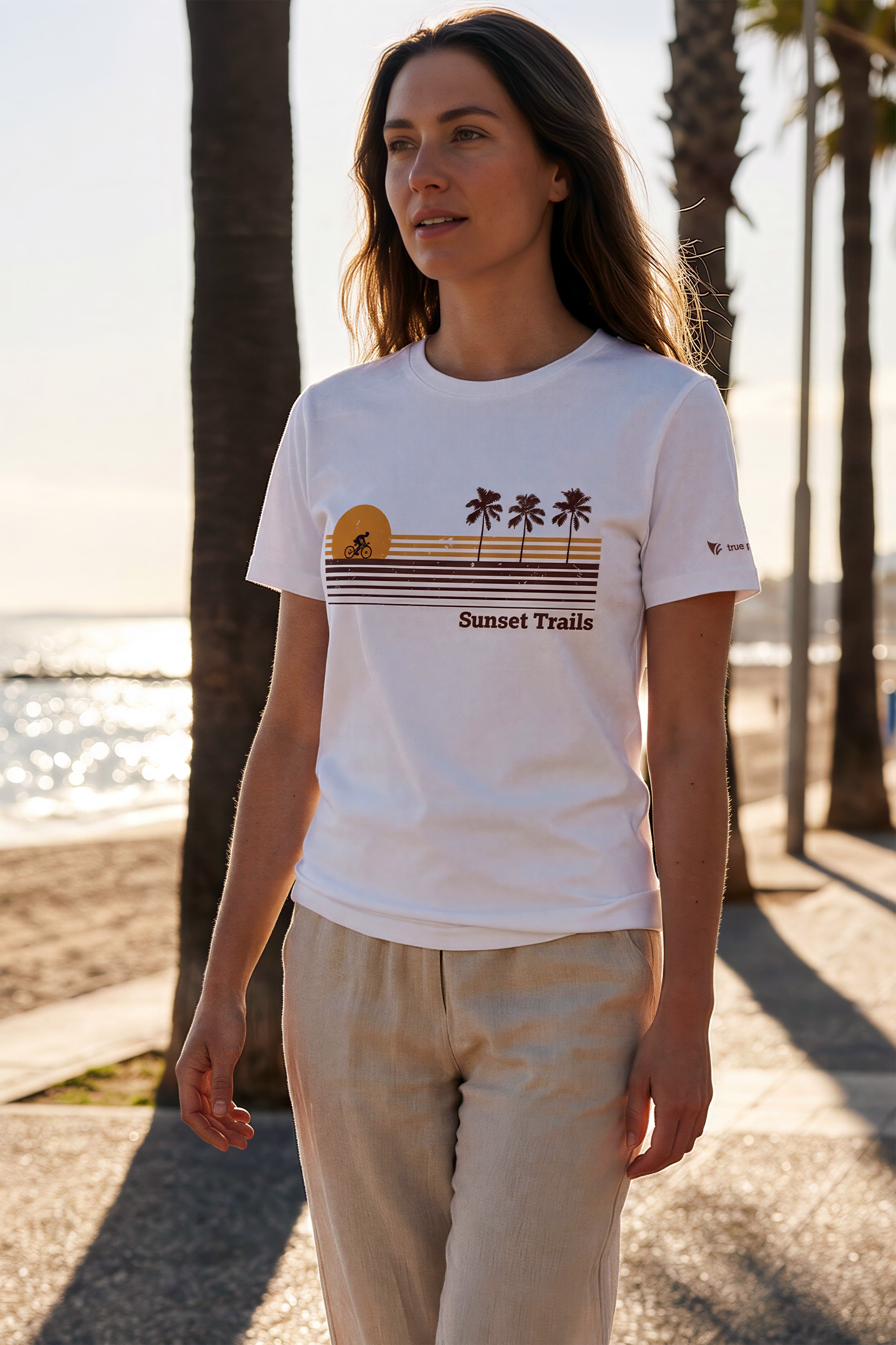 Woman wearing a white t-shirt with 'Sunset Trails' design by a beach with palm trees.