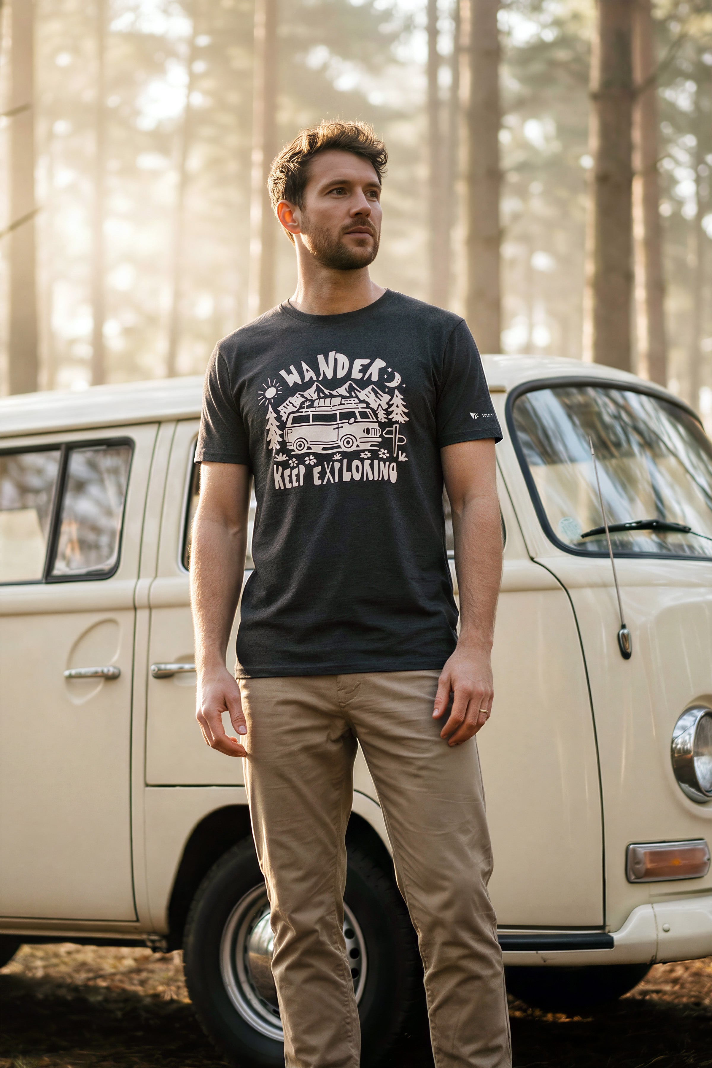 Man wearing a black t-shirt with a graphic design standing in front of a vintage van in a forest.