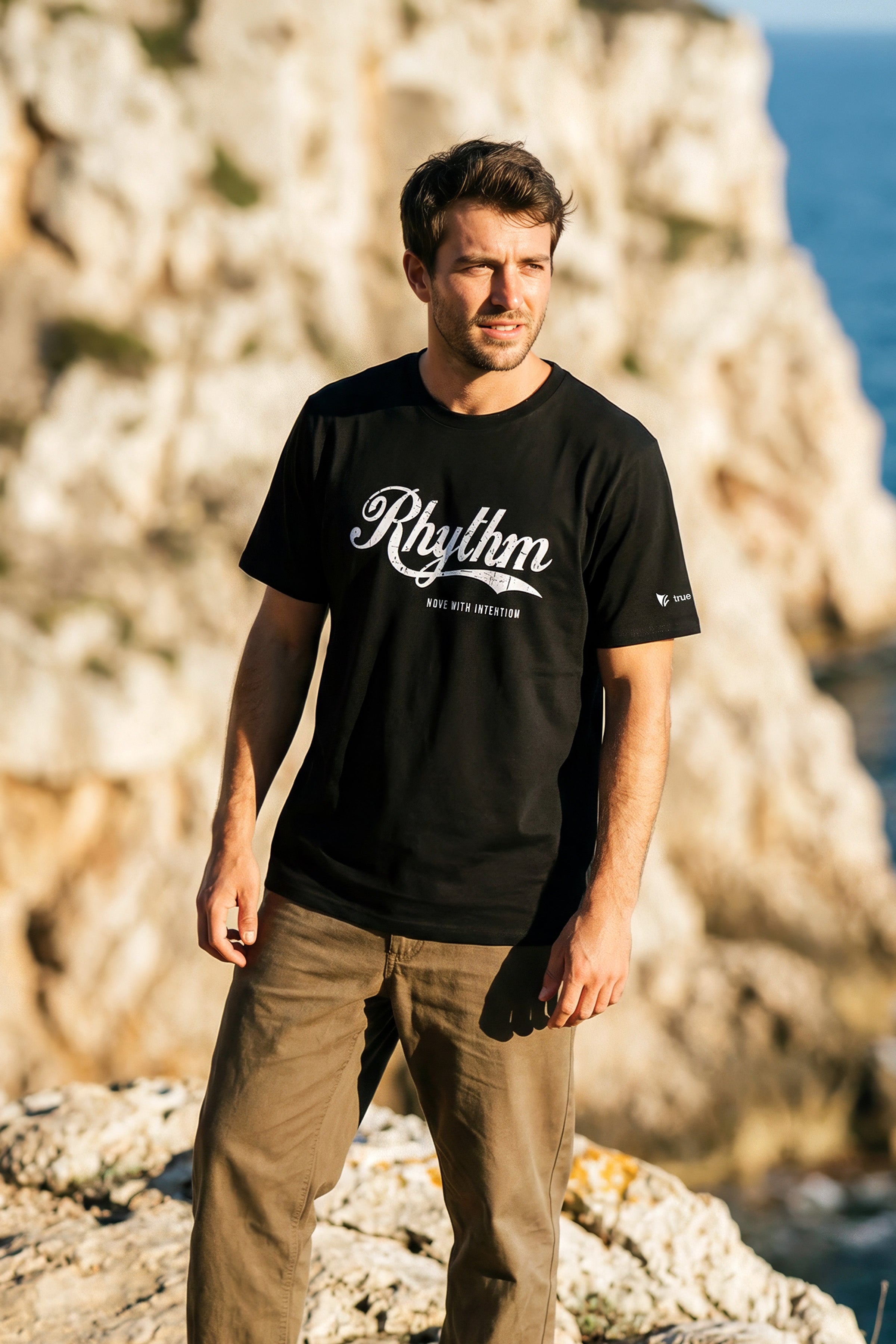 Man wearing a black t-shirt with 'Rhythm' logo standing on rocky terrain with ocean view