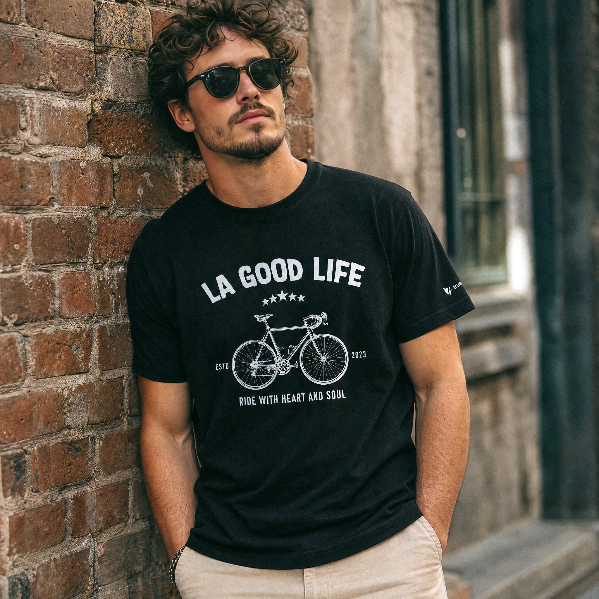 Man wearing a black t-shirt with 'LA GOOD LIFE' graphic leaning against a brick wall.