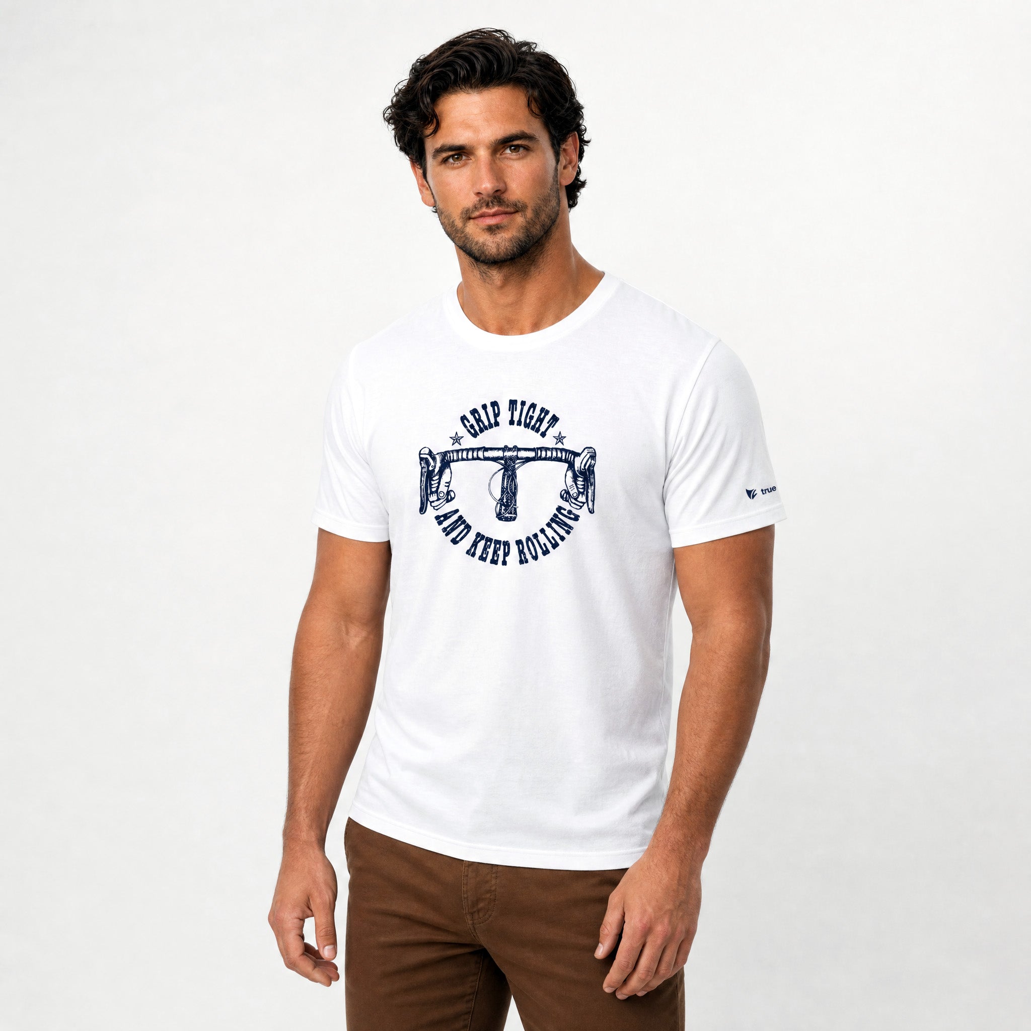 A man wearing a white crew-neck t-shirt with a navy blue graphic on the chest that reads 'CAMP FIGHT AND DEEP BOWLING', paired with brown pants, against a light gray studio background.