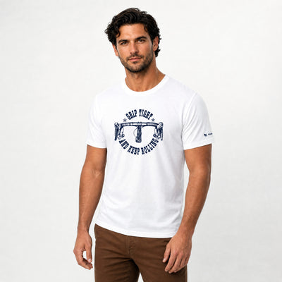 A man wearing a white crew-neck t-shirt with a navy blue graphic on the chest that reads 'CAMP FIGHT AND DEEP BOWLING', paired with brown pants, against a light gray studio background.